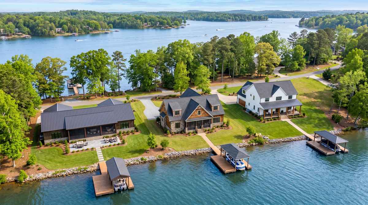 Aerial view of three newly built custom homes on adjacent waterfront lots along Lake Wylie, SC: a single-story modern ranch on the left with a low-pitched standing-seam roof, a 1.5-story craftsman with shed dormers and stone gable in the center, and a two-story modern farmhouse with board-and-batten siding on the right. Each home has a private covered dock extending into the lake, manicured lawns, mature hardwoods, and a gravel driveway, photographed in spring morning light